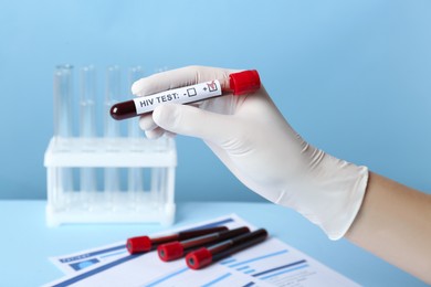 Photo of Scientist holding tube with blood sample and label HIV Test on light blue background, closeup