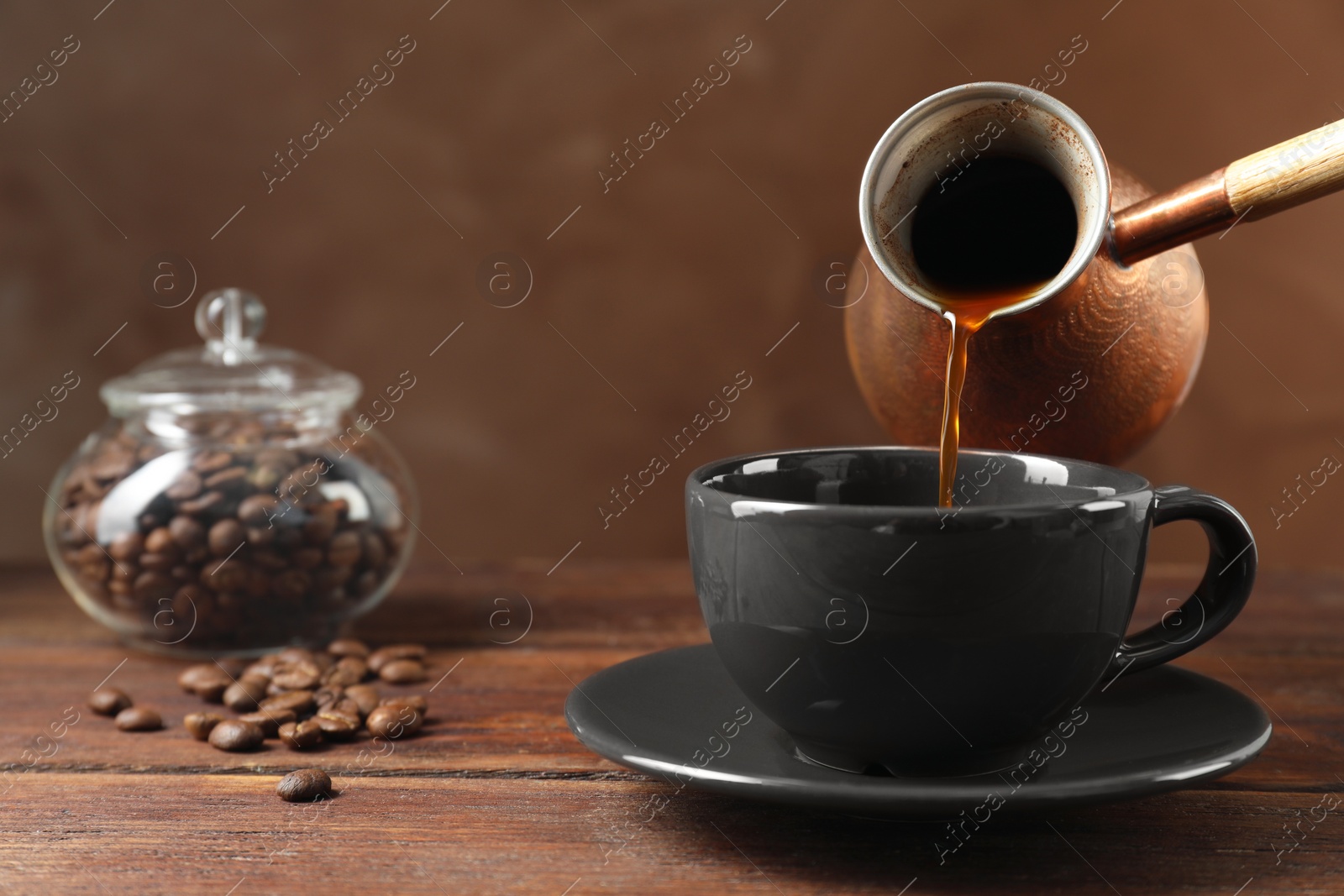 Photo of Turkish coffee. Pouring brewed beverage from cezve into cup at wooden table against brown background