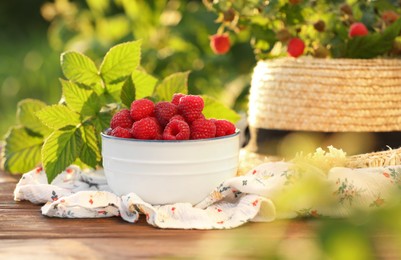 Photo of Tasty ripe raspberries in bowl, green leaves and straw hat on wooden table outdoors