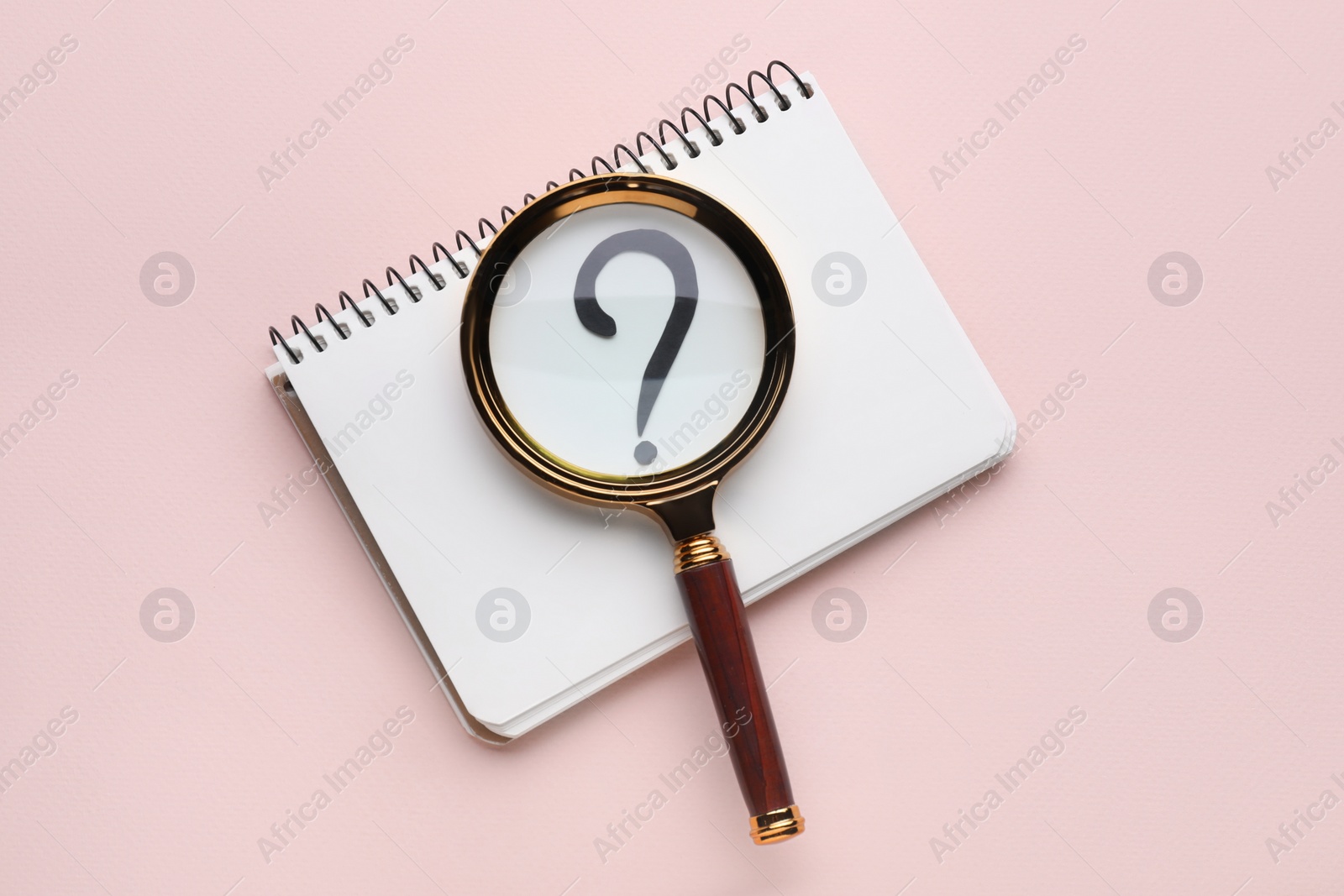 Photo of Magnifying glass over notebook with question mark on light pink background, top view
