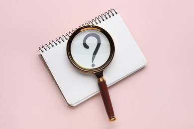 Magnifying glass over notebook with question mark on light pink background, top view