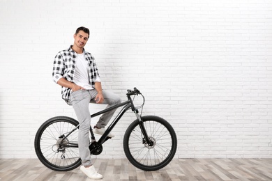 Photo of Handsome young man with modern bicycle near white brick wall indoors