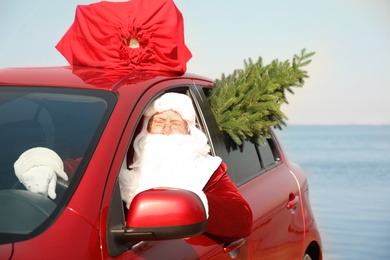 Photo of Authentic Santa Claus with fir tree and bag fullpresents on roof driving modern car  near sea