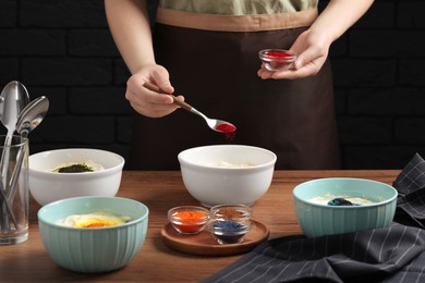 Photo of Woman adding red food coloring into bowl at wooden table, closeup
