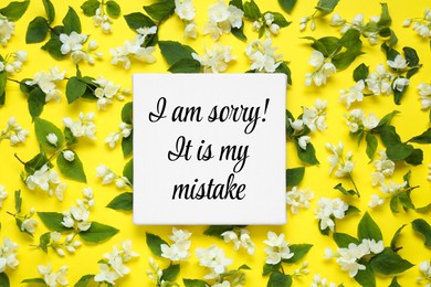 Canvas with sincere sorry message and many jasmine flowers on yellow background, flat lay