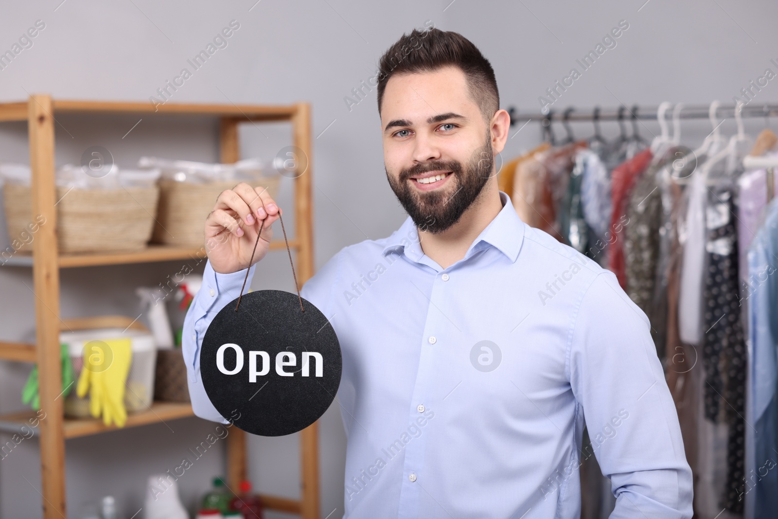 Photo of Dry-cleaning service. Happy worker holding Open sign indoors