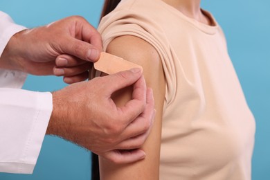 Doctor sticking plaster on woman's arm after vaccination against light blue background, closeup