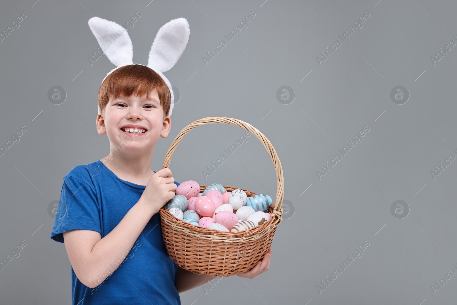 Photo of Easter celebration. Cute little boy with bunny ears and wicker basket full of painted eggs on grey background. Space for text