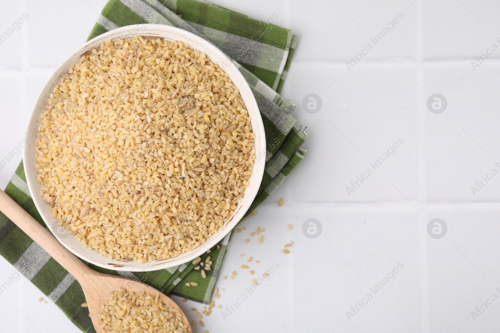 Photo of Bowl and spoon with raw bulgur on white tiled table, top view. Space for text