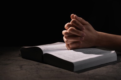 Religion. Christian woman praying over Bible at table against black background, closeup. Space for text