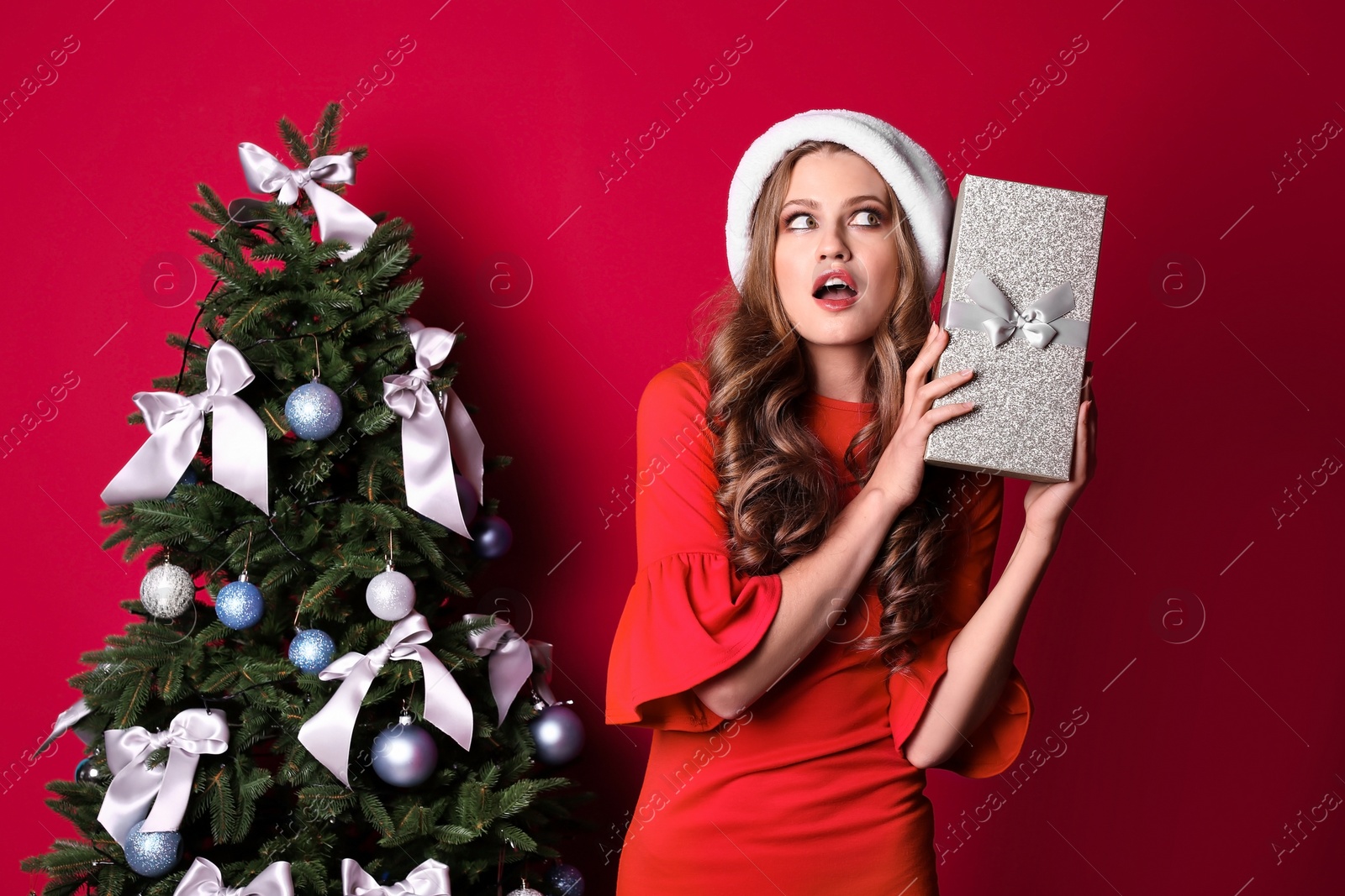 Photo of Beautiful young woman in Santa hat with gift box near Christmas tree on color background