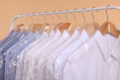 Photo of Dry-cleaning service. Many different clothes in plastic bags hanging on rack against beige background, closeup