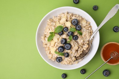 Photo of Tasty oatmeal with blueberries, mint and almond petals in bowl on light green background, flat lay. Space for text