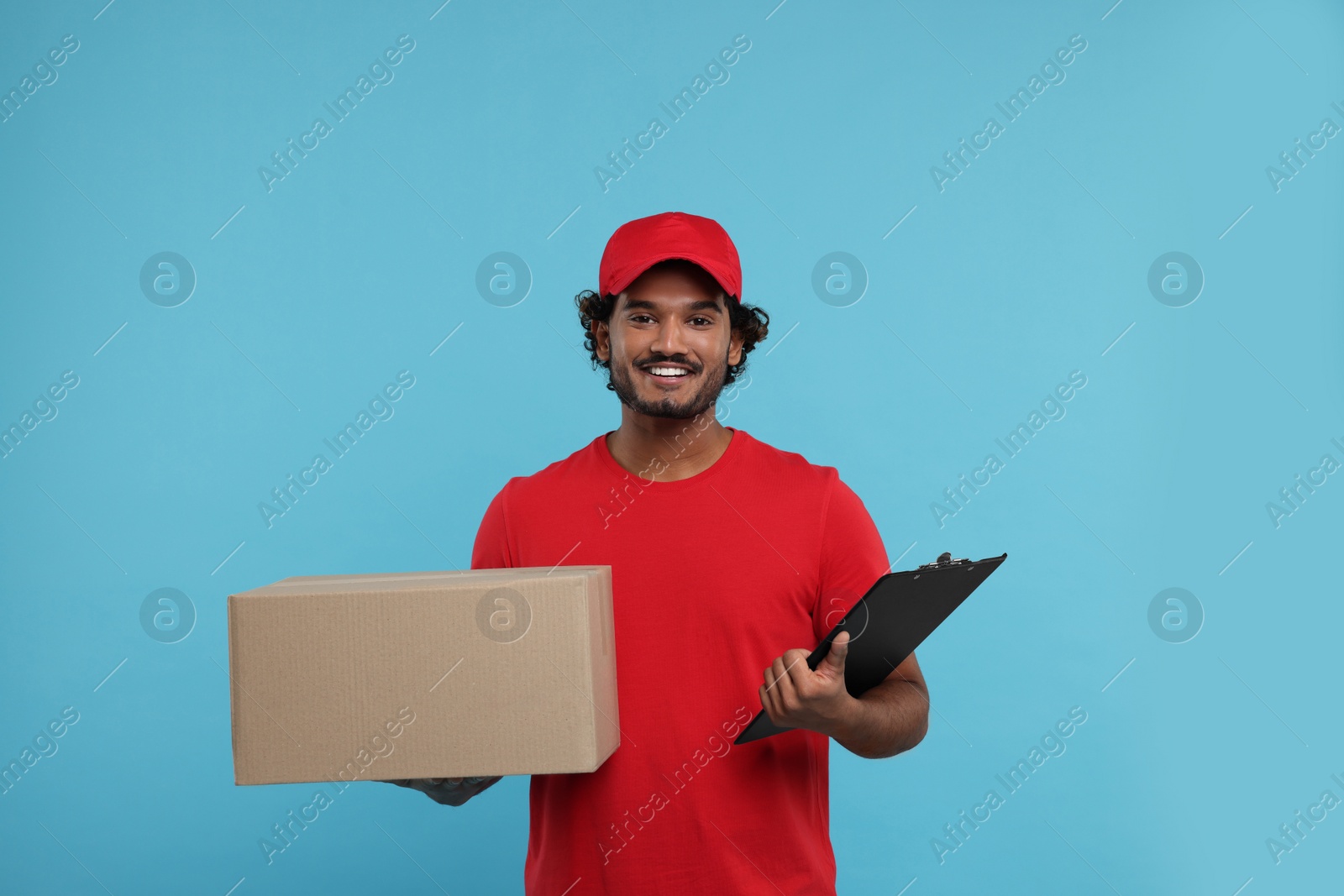 Photo of Happy young courier with parcel and clipboard on light blue background