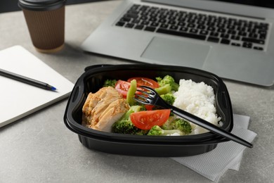 Container with tasty food, laptop, fork and notebook on light grey table. Business lunch