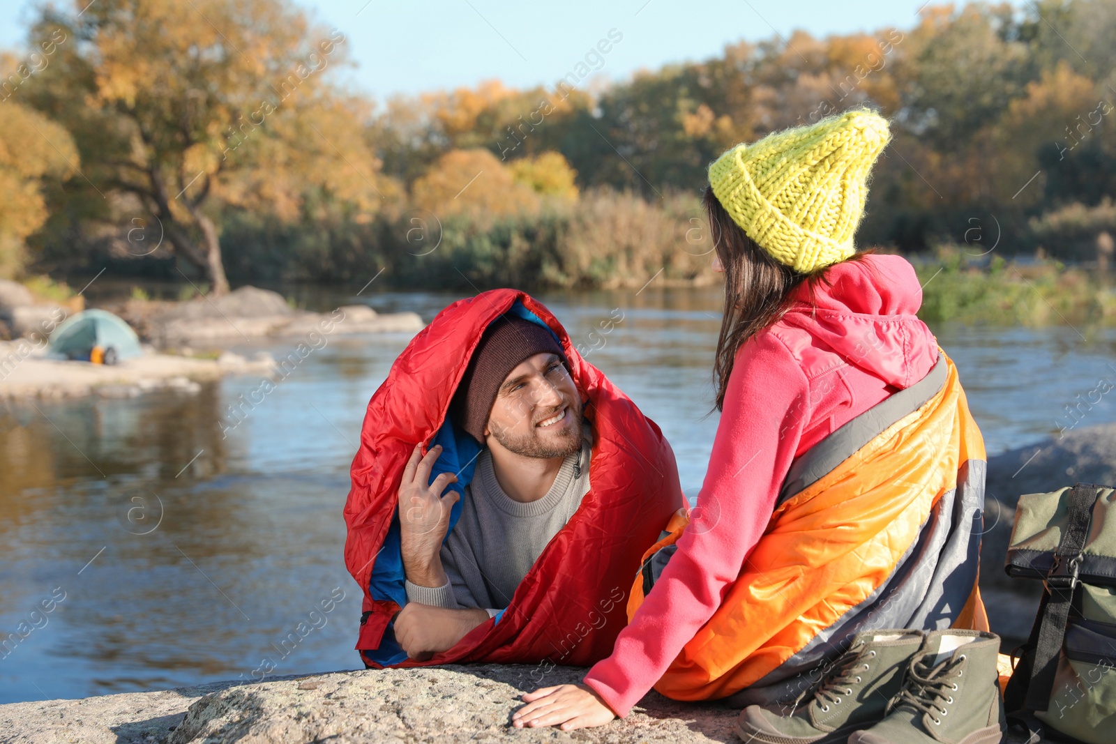 Photo of Couple of campers in sleeping bags sitting on rock near pond. Space for text