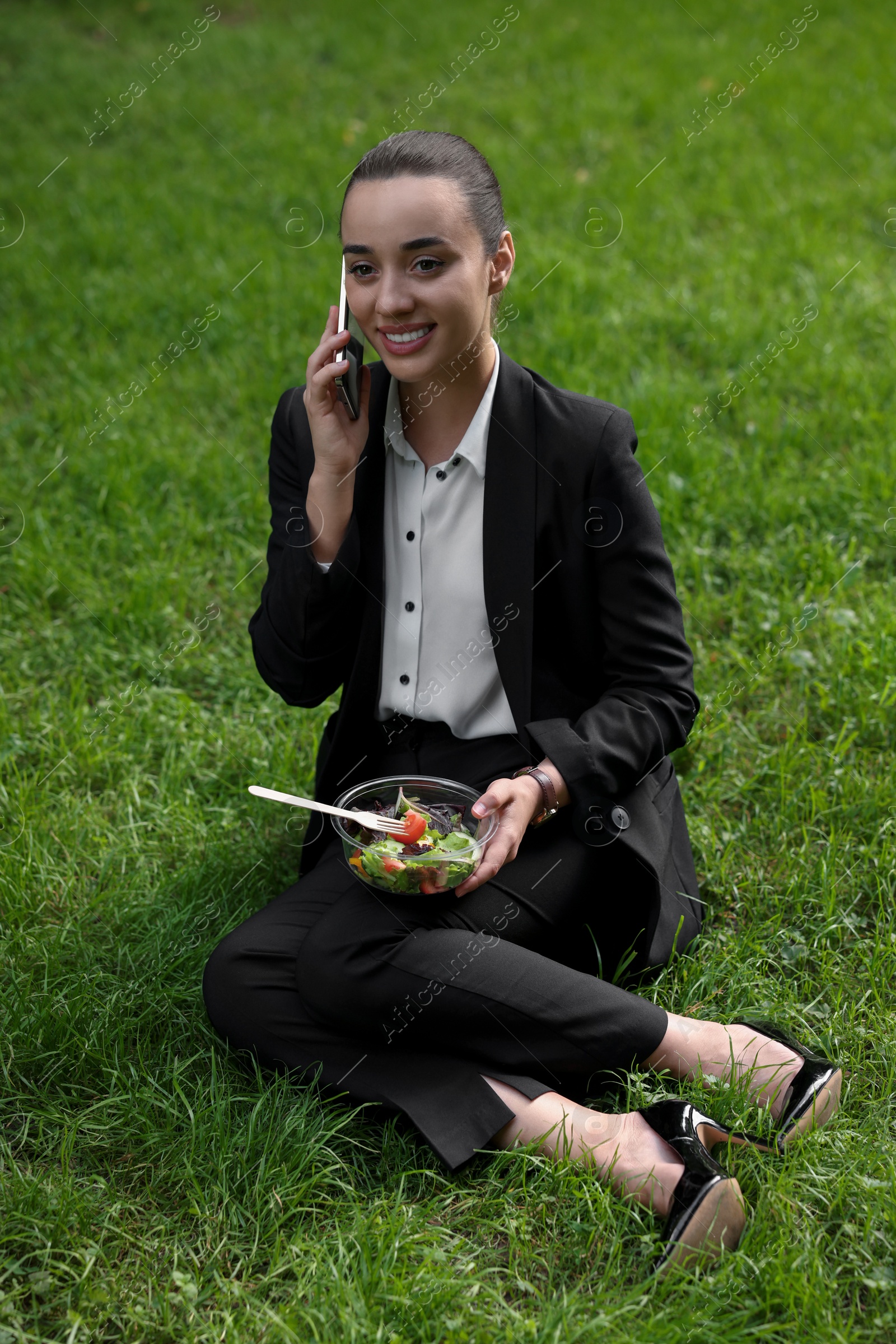 Photo of Lunch time. Happy businesswoman with container of salad talking on smartphone on green grass outdoors