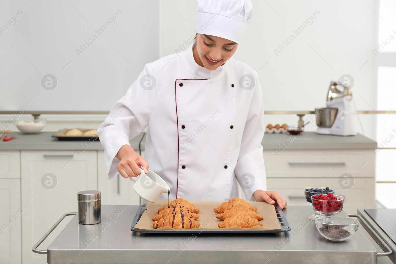 Photo of Young female pastry chef pouring chocolate sauce onto croissants at table in kitchen