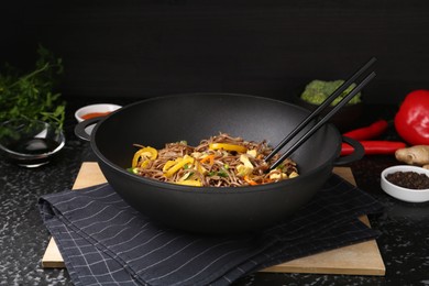 Photo of Stir-fry. Tasty noodles with vegetables, meat and chopsticks in wok on dark textured table