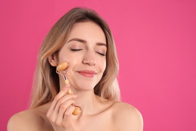 Young woman using natural jade face roller on pink background