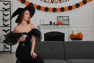 Young woman in scary witch costume with glass of cocktail indoors, space for text. Halloween celebration