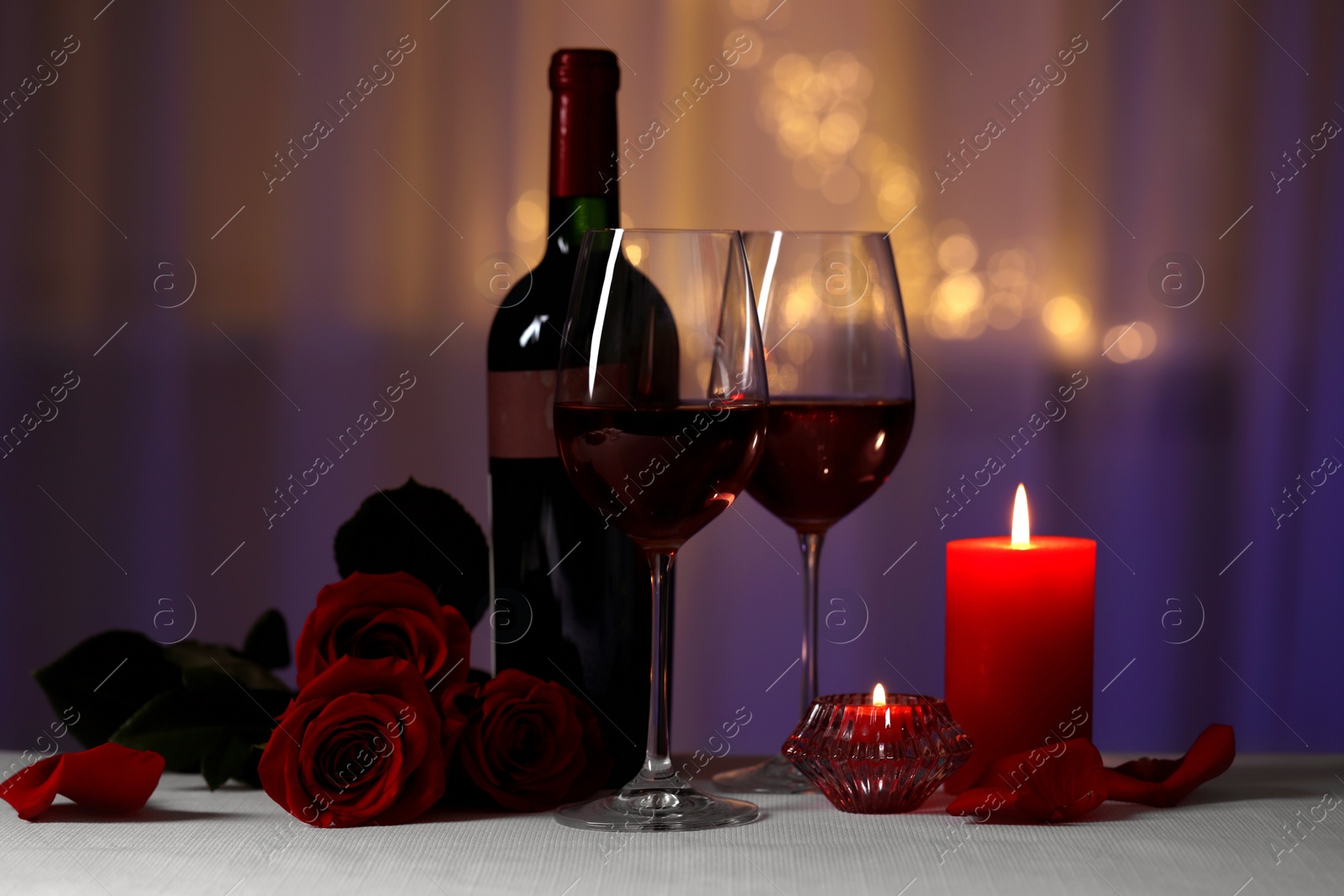 Photo of Glasses of red wine, rose flowers and burning candles on grey table against blurred lights. Romantic atmosphere