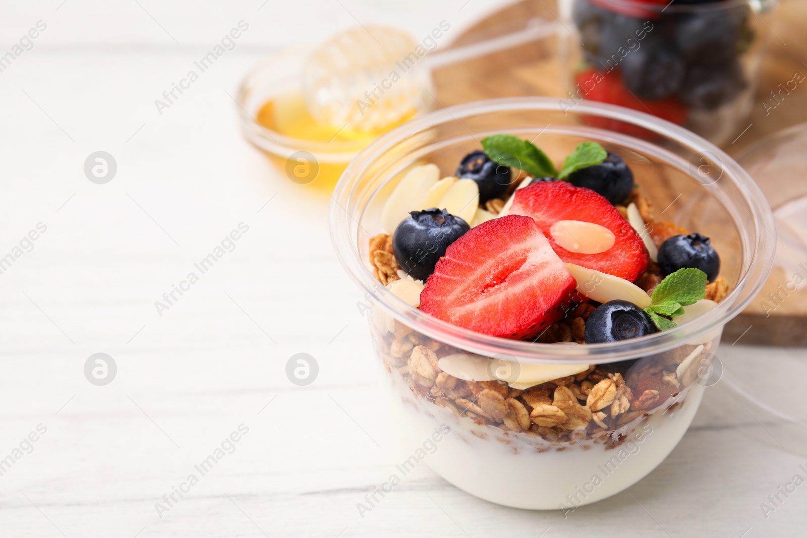 Photo of Tasty granola with berries, yogurt and almond flakes in plastic cup on white wooden table, closeup. Space for text