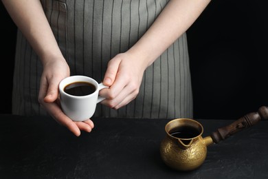 Photo of Turkish coffee. Woman with cup of freshly brewed beverage and cezve at black table, closeup