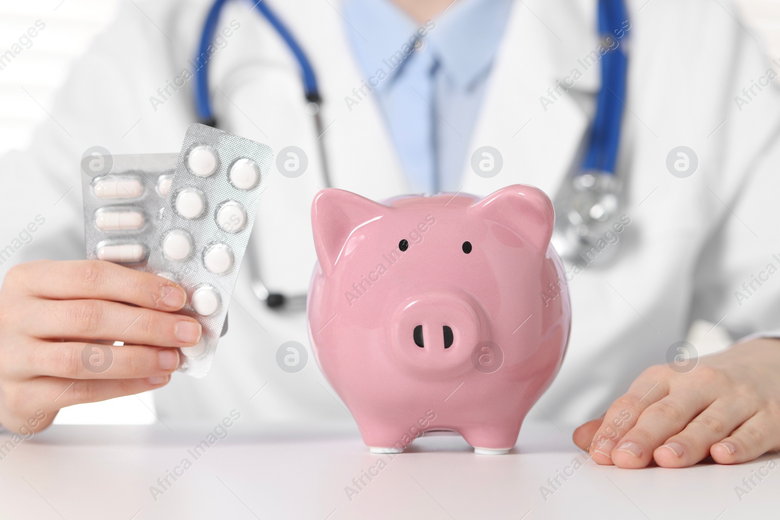 Photo of Doctor with pills and piggy bank at white table, closeup