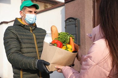 Photo of Courier in medical mask giving paper bag with groceries to woman outdoors. Delivery service Covid-19 during quarantine