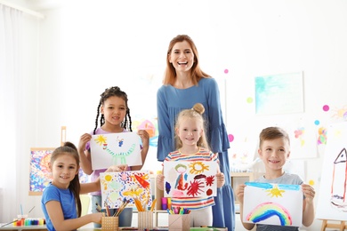 Photo of Cute little children with teacher showing their paintings at lesson indoors