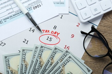 Photo of Calendar with date reminder about tax day, documents, pen, money, glasses and calculator on wooden table, flat lay