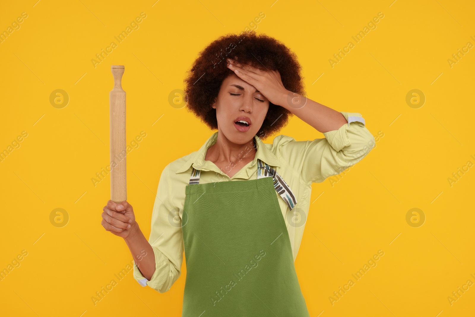 Photo of Frustrated young woman in apron holding rolling pin on orange background