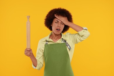 Photo of Frustrated young woman in apron holding rolling pin on orange background