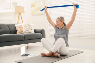 Photo of Senior woman doing exercise with fitness elastic band near laptop on mat at home