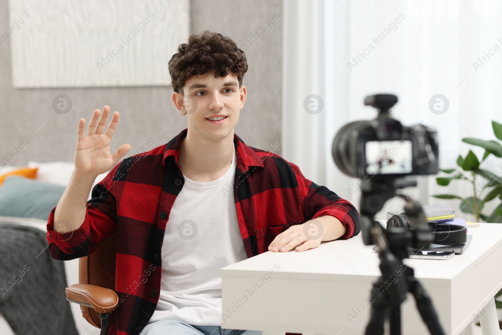 Photo of Smiling teenage blogger waving hello to his subscribers while streaming at home