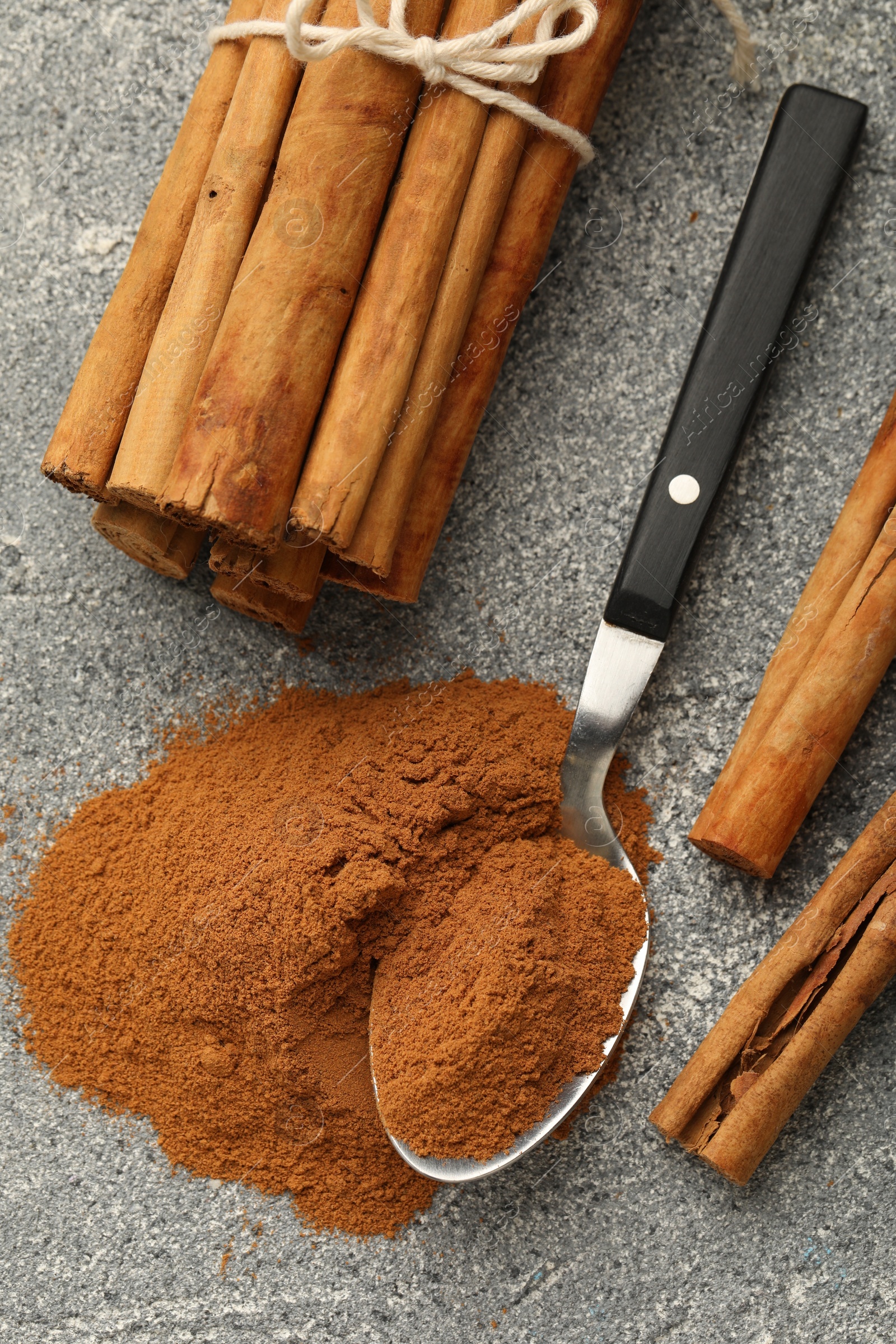 Photo of Spoon with cinnamon powder and sticks on grey table, flat lay