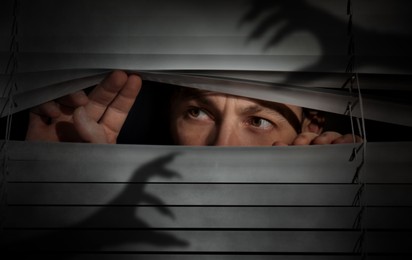 Image of Worried man looking through window blinds into darkness. Shadow of hands with long claws. Paranoia concept