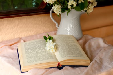 Photo of Open book and beautiful jasmine flowers on pink fabric indoors