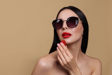 Photo of Attractive woman in fashionable sunglasses holding strawberry against beige background. Space for text