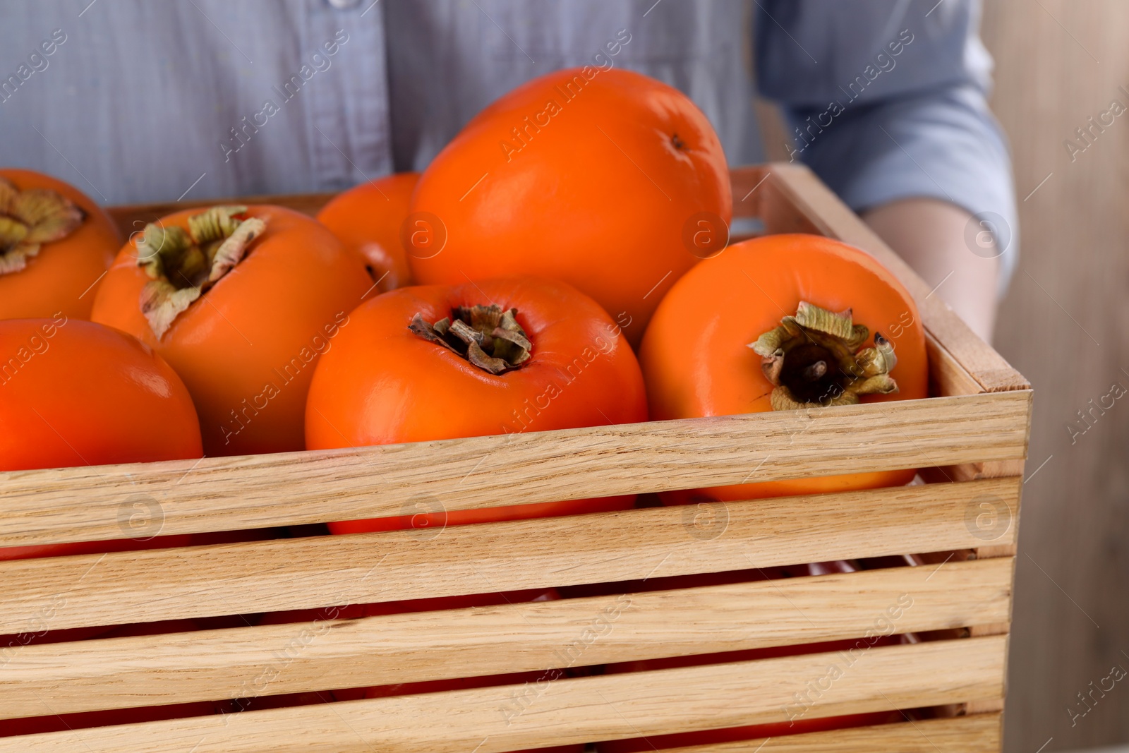 Photo of Woman holding wooden crate with delicious ripe juicy persimmons on blurred background, closeup