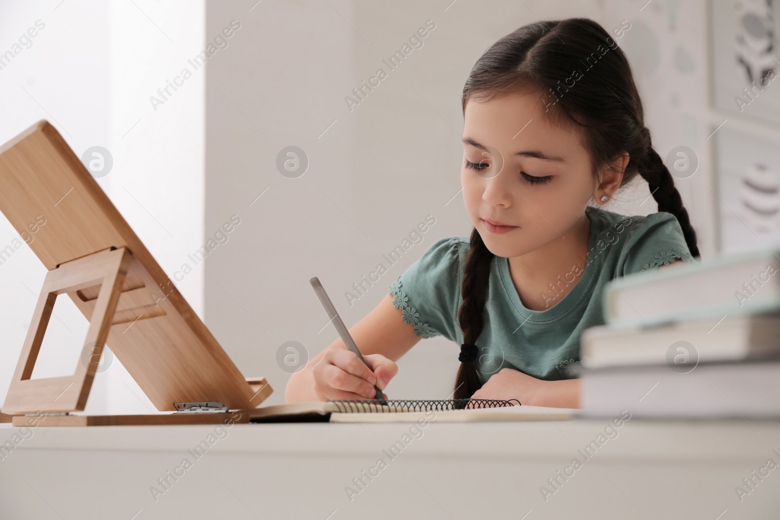 Photo of Little girl doing homework with tablet at table in room