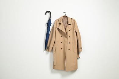 Photo of Hanger with beige trench coat and blue umbrella on white wall, space for text