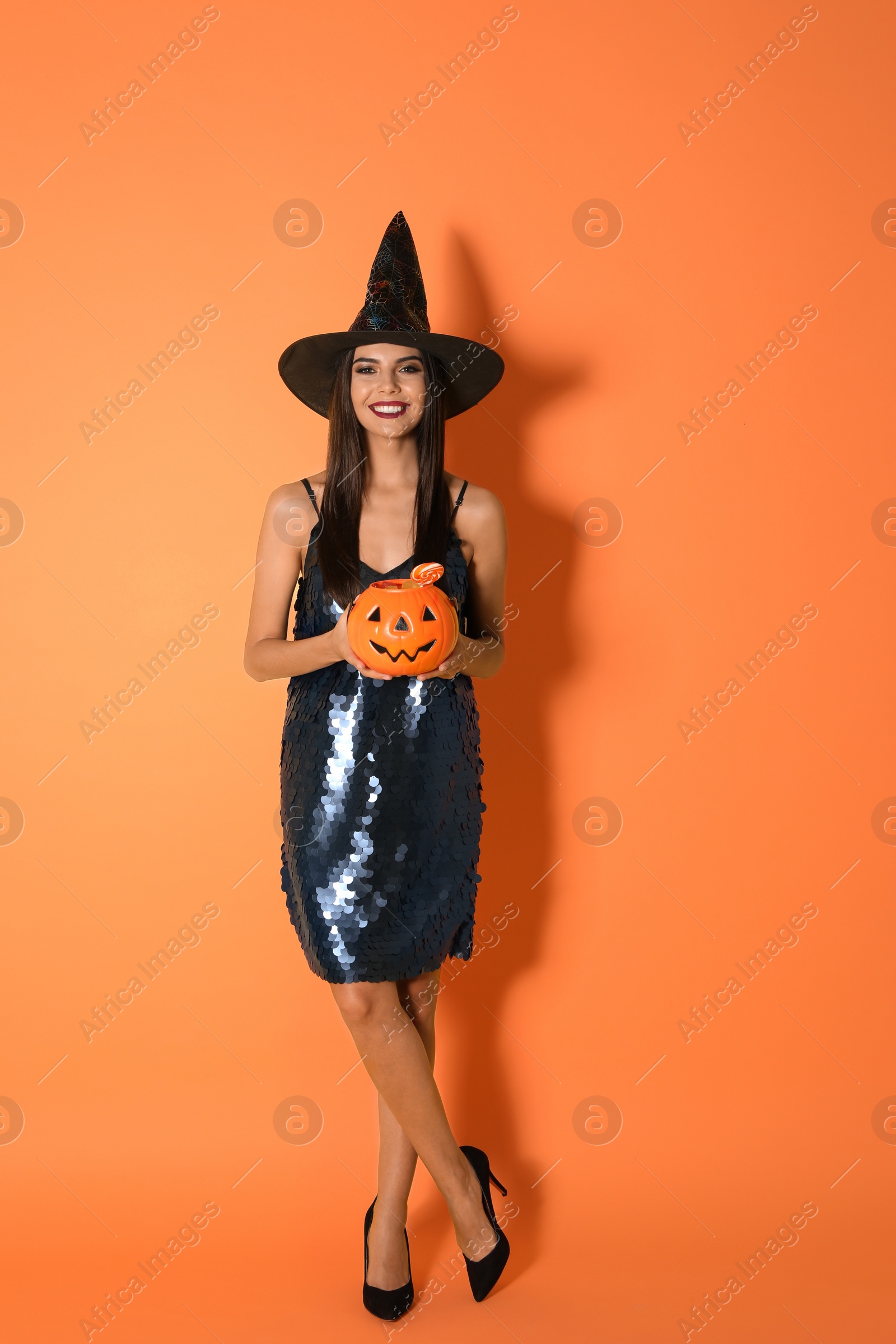 Photo of Beautiful woman wearing witch costume with Jack O'Lantern candy container for Halloween party on yellow background