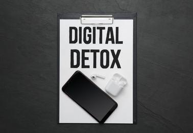 Photo of Clipboard with phrase DIGITAL DETOX, smartphone and earphones on black stone table, flat lay