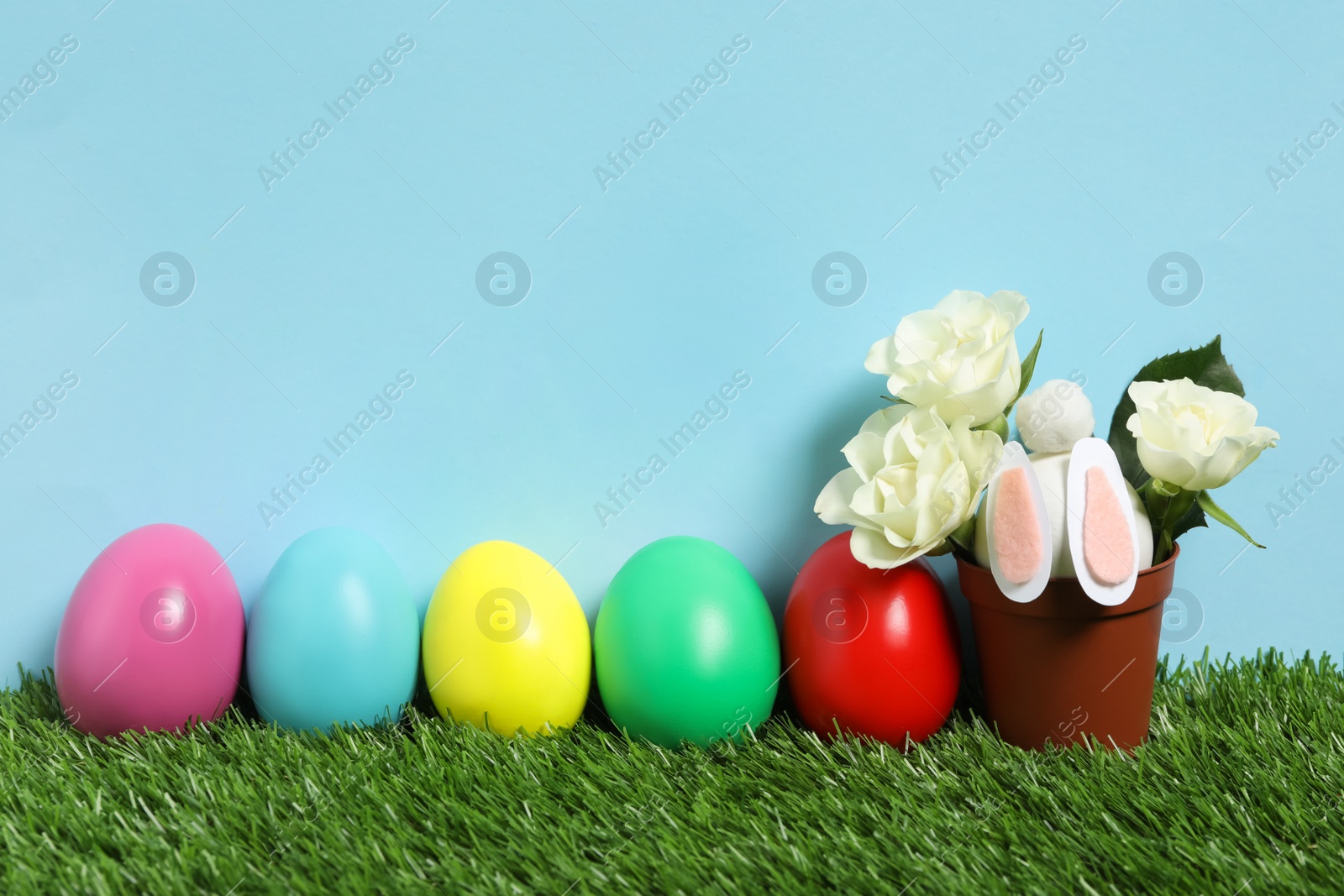 Photo of Bright Easter eggs, pot with flowers and toy bunny on green grass against light blue background