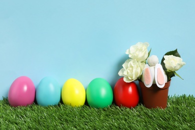Photo of Bright Easter eggs, pot with flowers and toy bunny on green grass against light blue background
