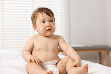 Cute little baby with moisturizing cream on face sitting on bed indoors