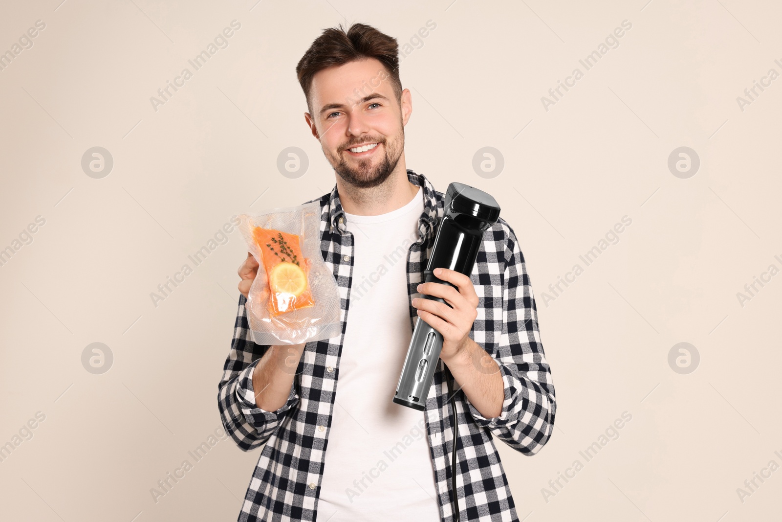 Photo of Smiling man holding sous vide cooker and salmon in vacuum pack on beige background