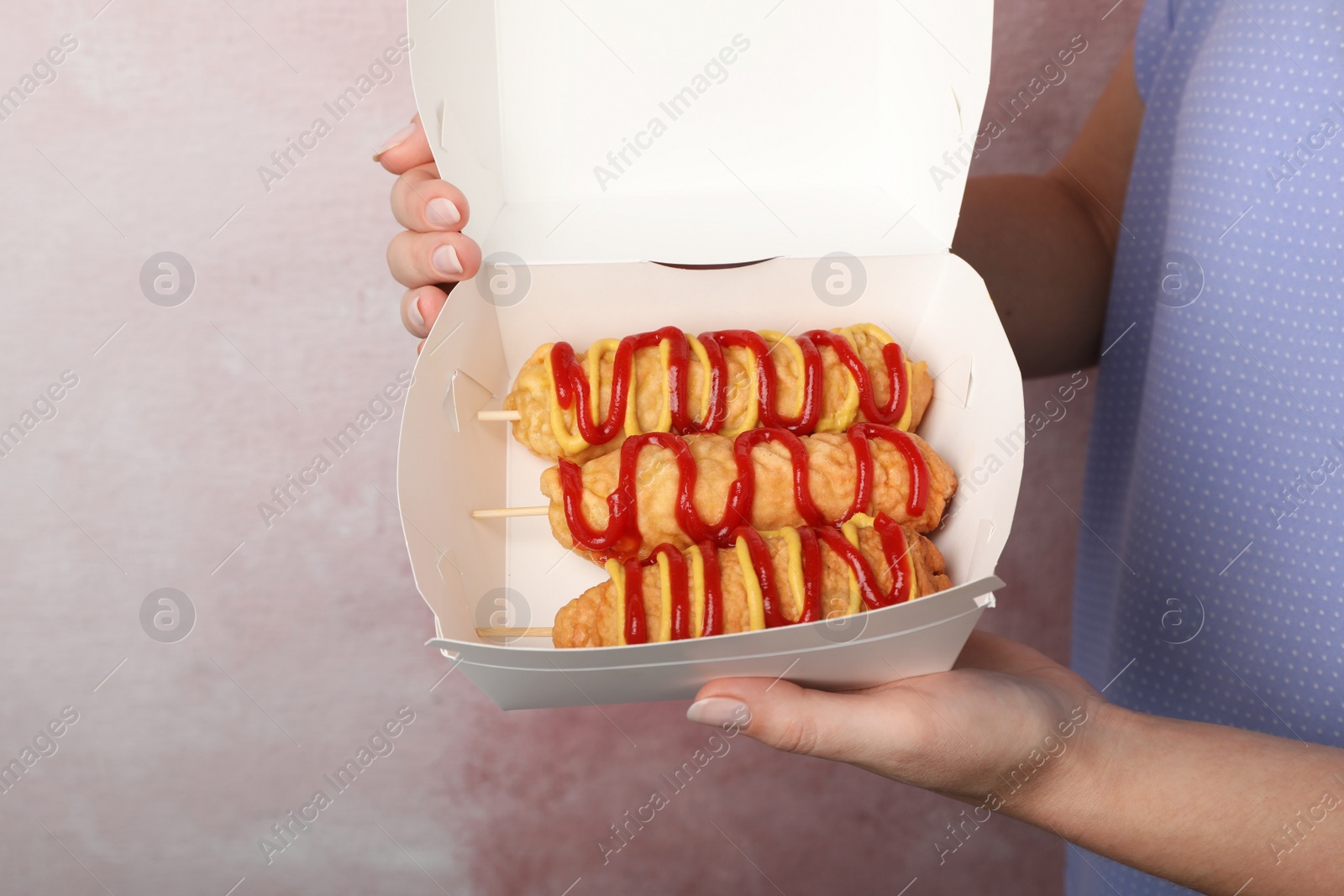 Photo of Woman holding box of delicious corn dogs with mustard and ketchup on pink background, closeup